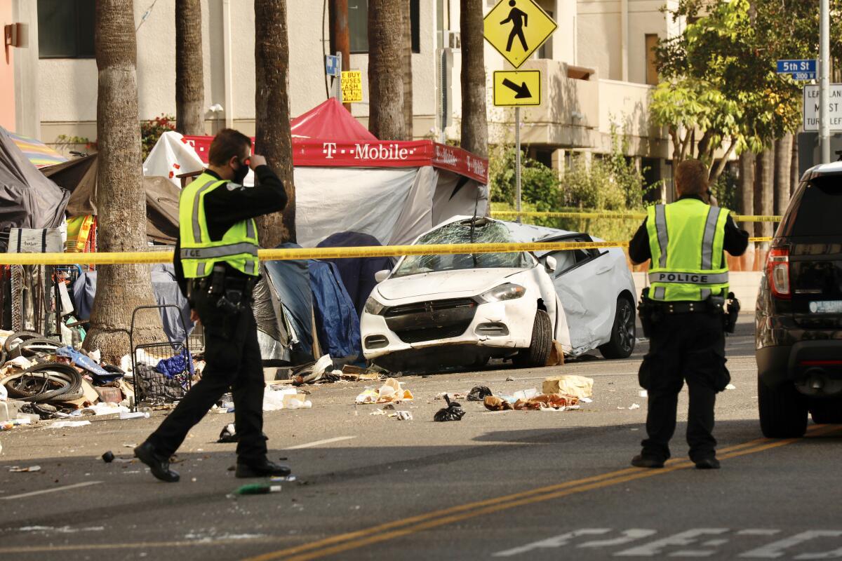 Two men in dark police uniform stand near yellow police tape cordoning off a wrecked white car near buildings 