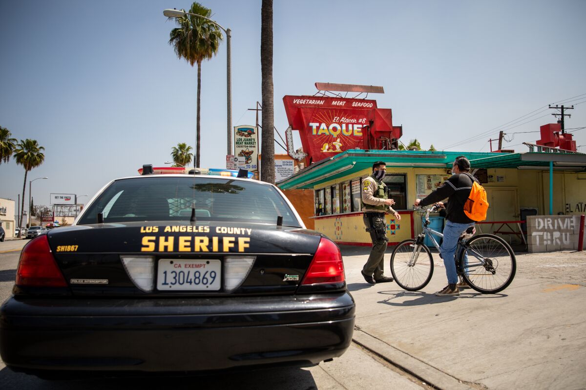 A sheriff's deputy speaks to a bicyclist on the sidewalk near his squad car and a taco stand