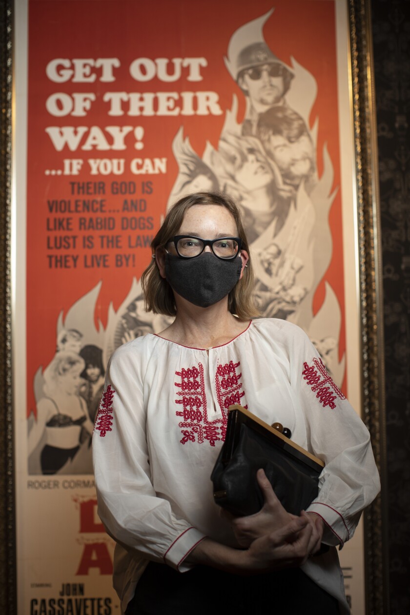 A woman wearing a dark mask and holding a dark purse stands in front of a movie poster.
