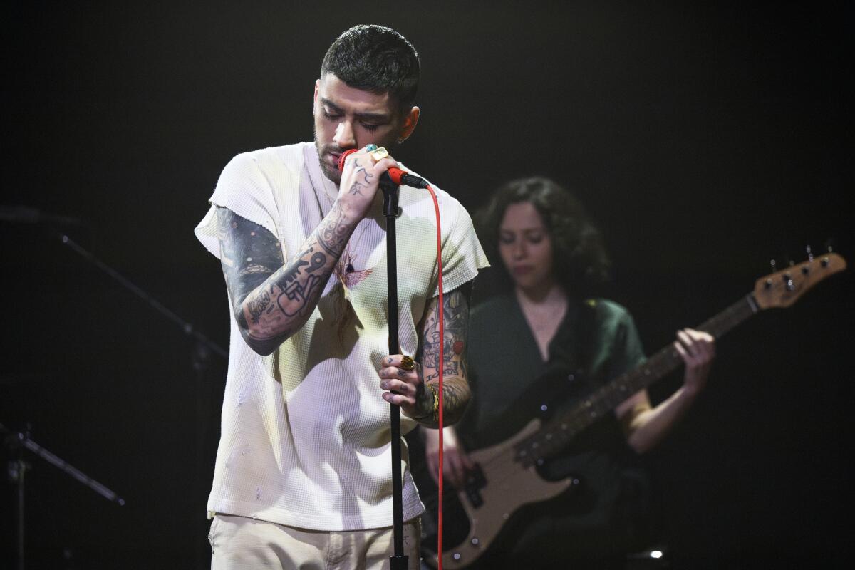 A man in a T-shirt sings into a microphone on a stand while a woman plays guitar behind him