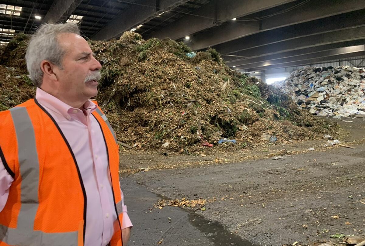 A man wearing an orange reflective vest stands before a pile of refuse.