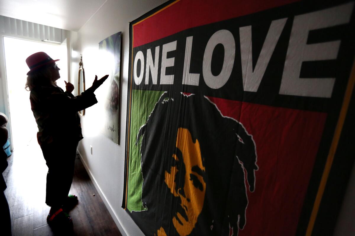 A woman is silhouetted while standing beside an illustration of Bob Marley on a rasta-colored tapestry that reads "One Love"
