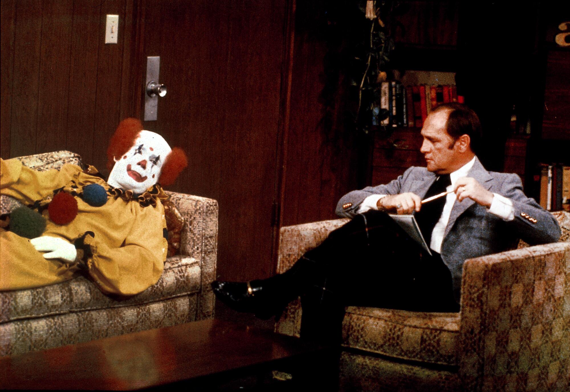 Bob Newhart sits in a chair with a fully costumed clown lying on the couch next to him