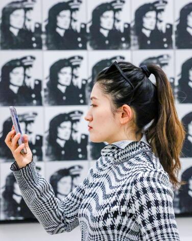 A woman in black and white houndstooth takes a selfie in front of a black-and-white photo grid hanging on the wall