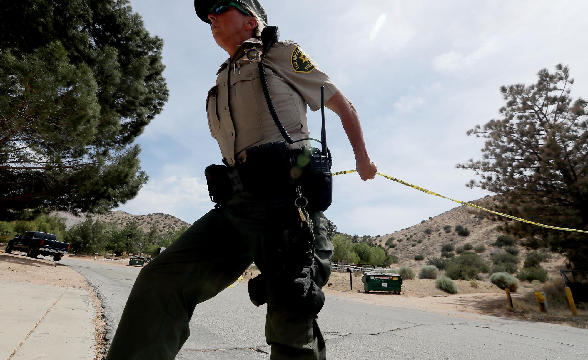 A deputy stretches police tape across a street surrounded by low, scrub-covered hills.