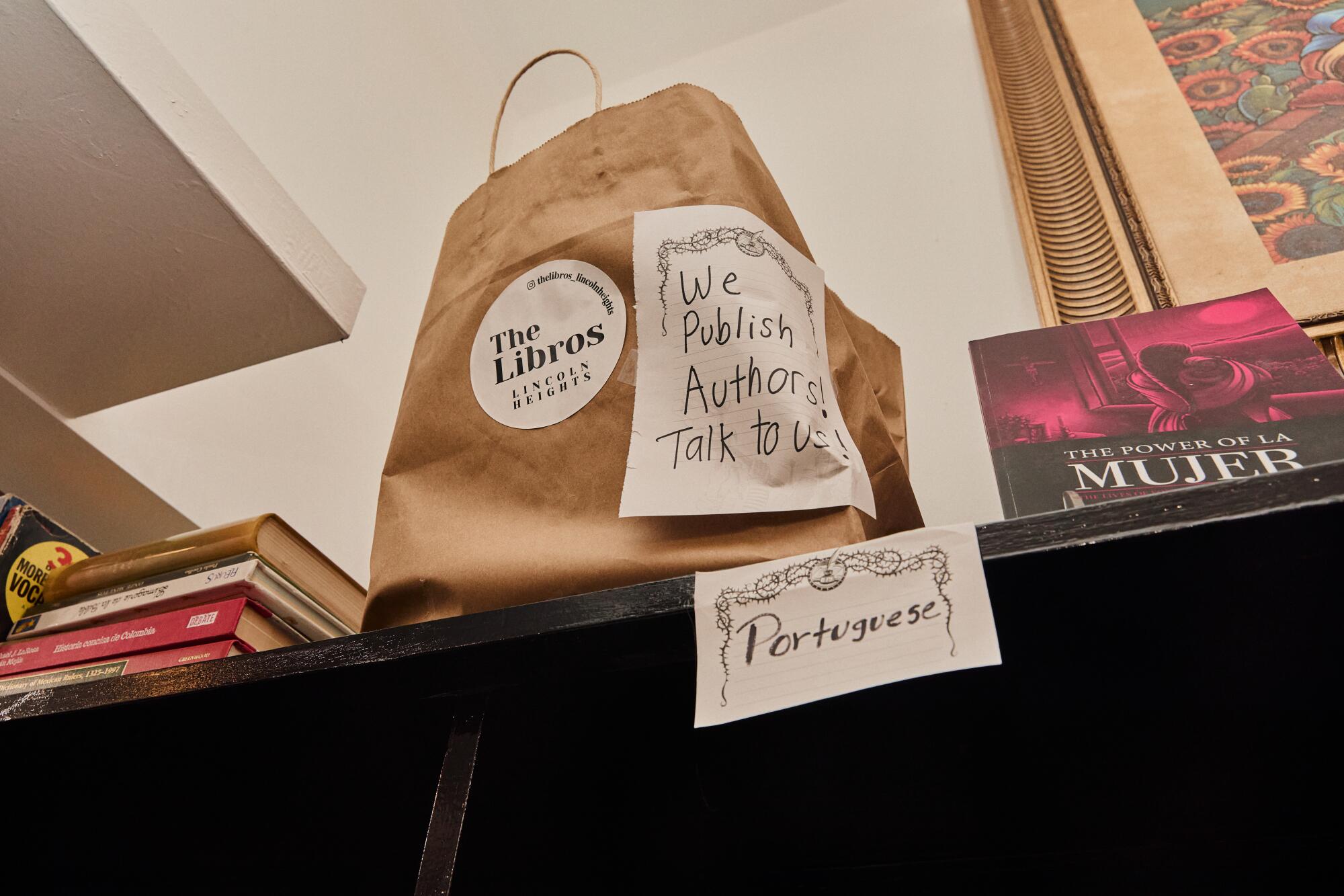 A paper bag with a sign that says "We publish authors, talk to us" It is located on top of a shelf in the Libros bookstore.