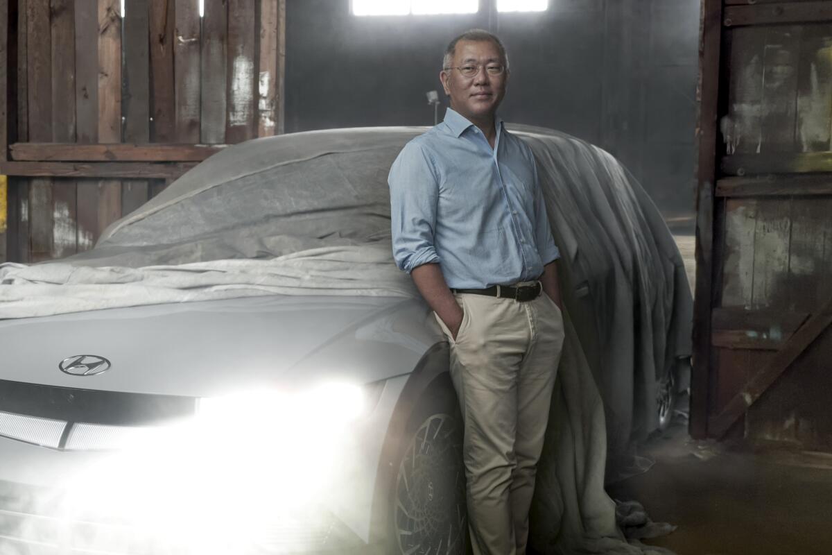 A man in a blue shirt and khaki pants stands next to a white Hyundai with its headlights on. 