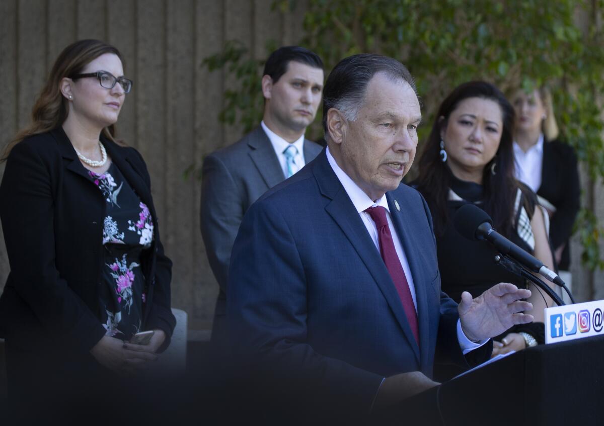 A man in a suit and tie speaks as other people look on.