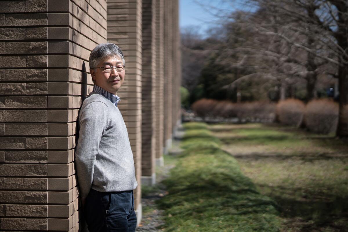 Yushi Inaba poses for a portrait in a sunny spot, leaning against a yellow-brick wall.