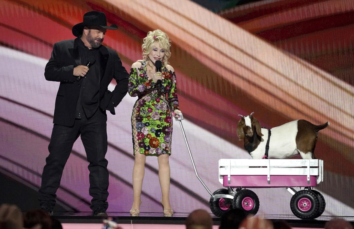 Garth Brooks, left, and Dolly Parton hold microphones on stage with a goat in a pink and white wheeled wagon with a handle