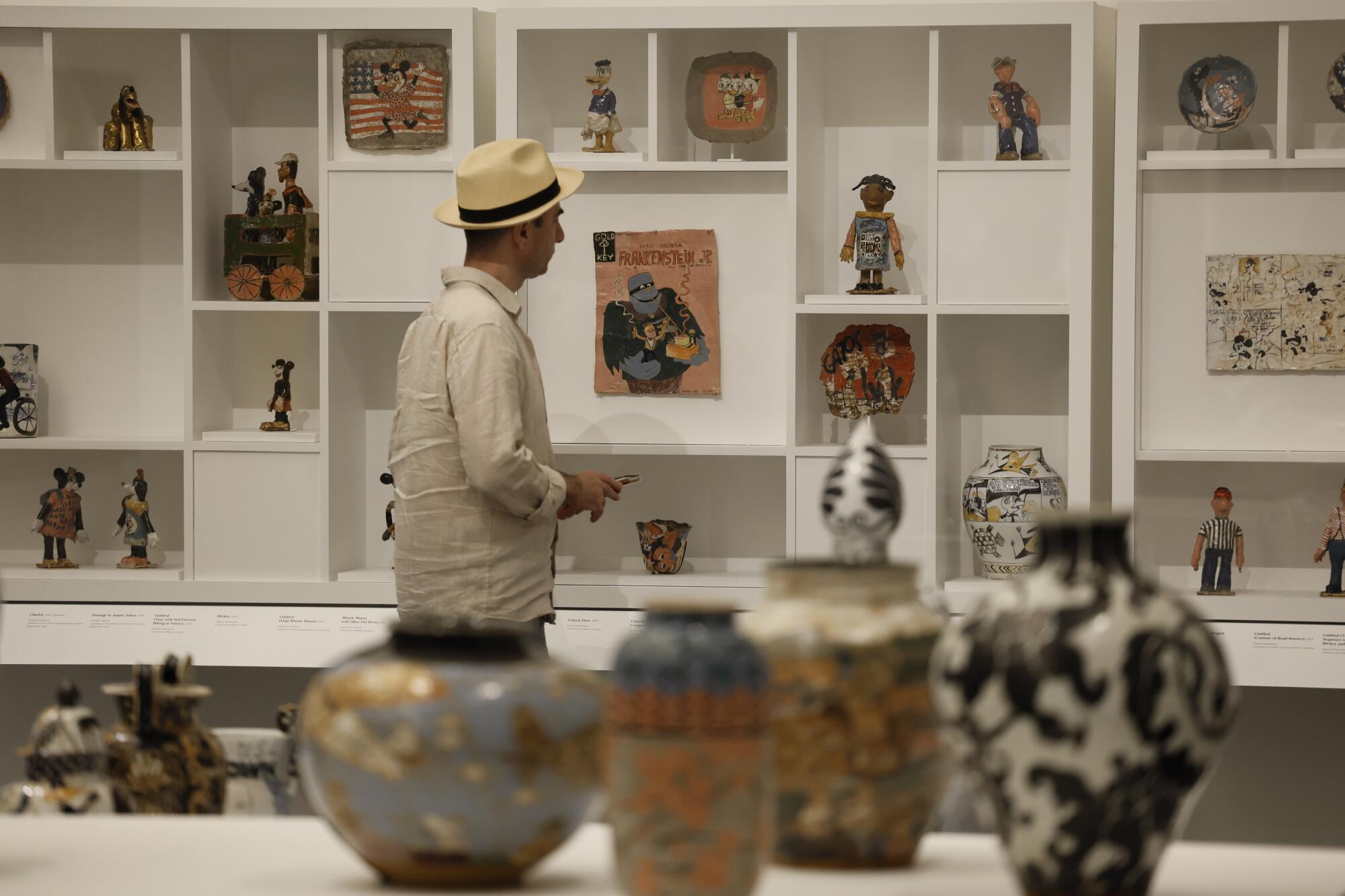 A man walks through a gallery, its white wall lined with  small-scale sculptures and other painted ceramics.