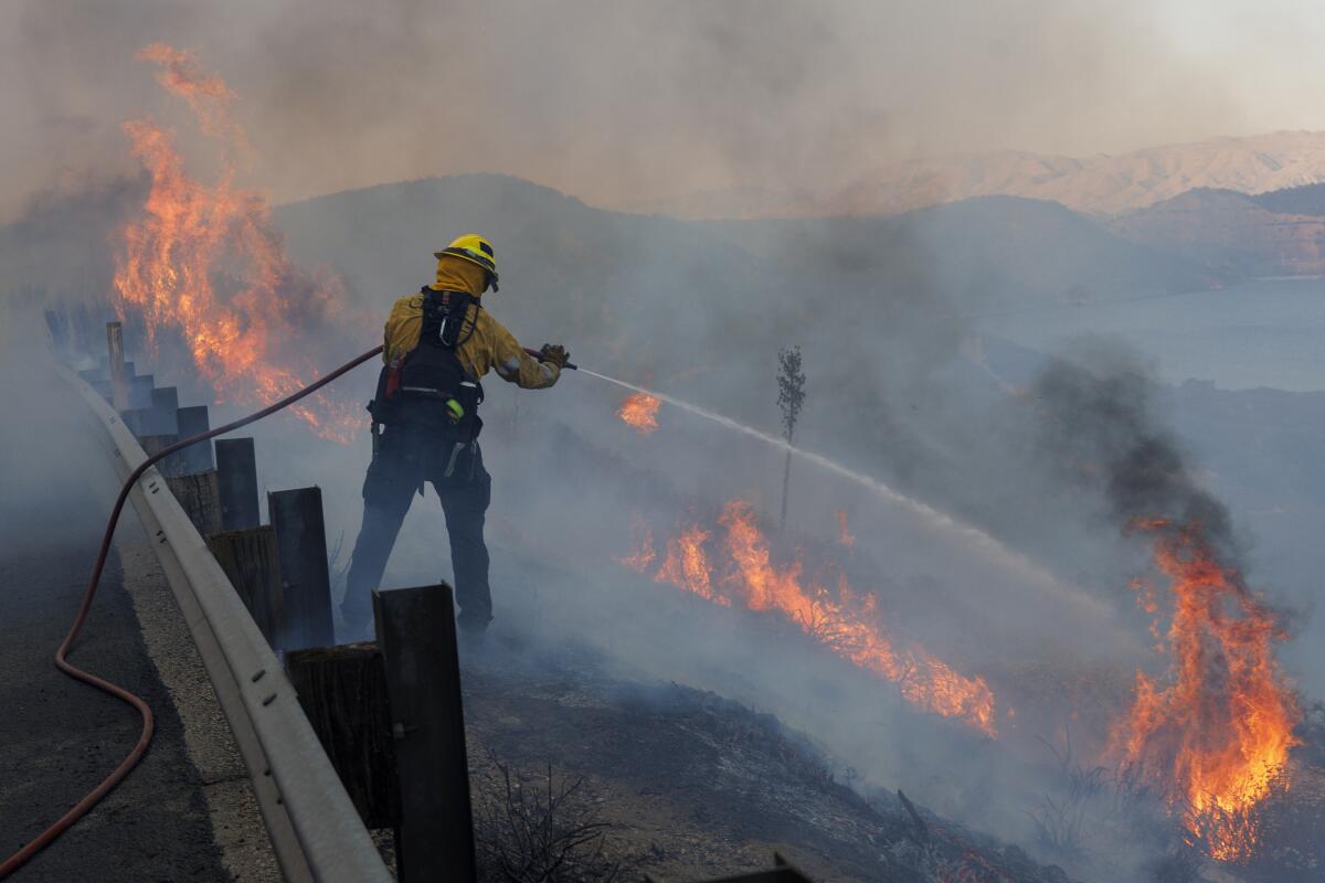A Cal Fire firefighter points a hose at flames from a brush fire off the highway.