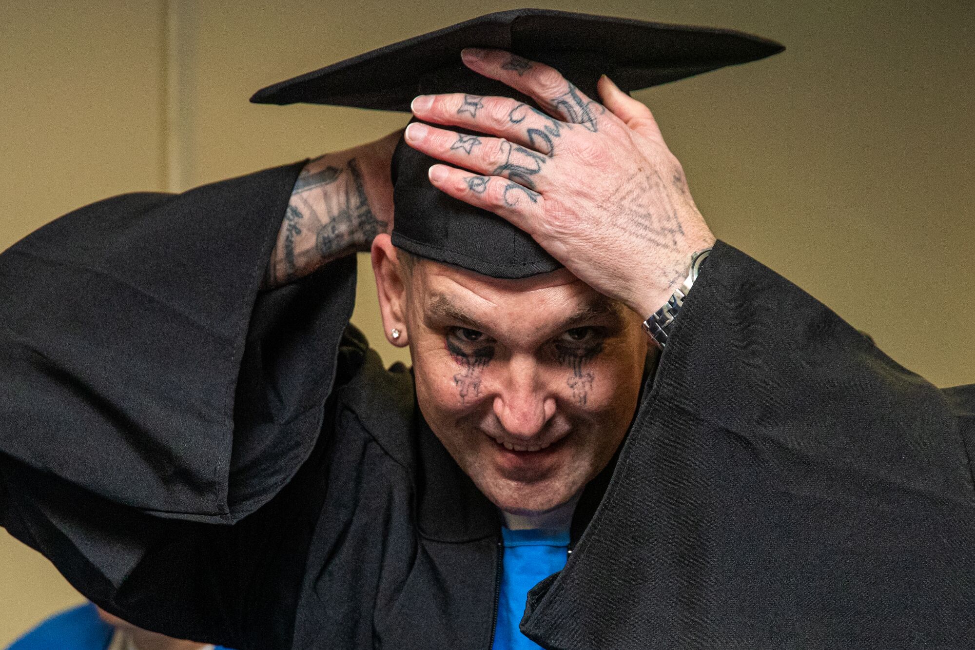A man with tattoos on his hands and face adjusts the cap of his cap and gown