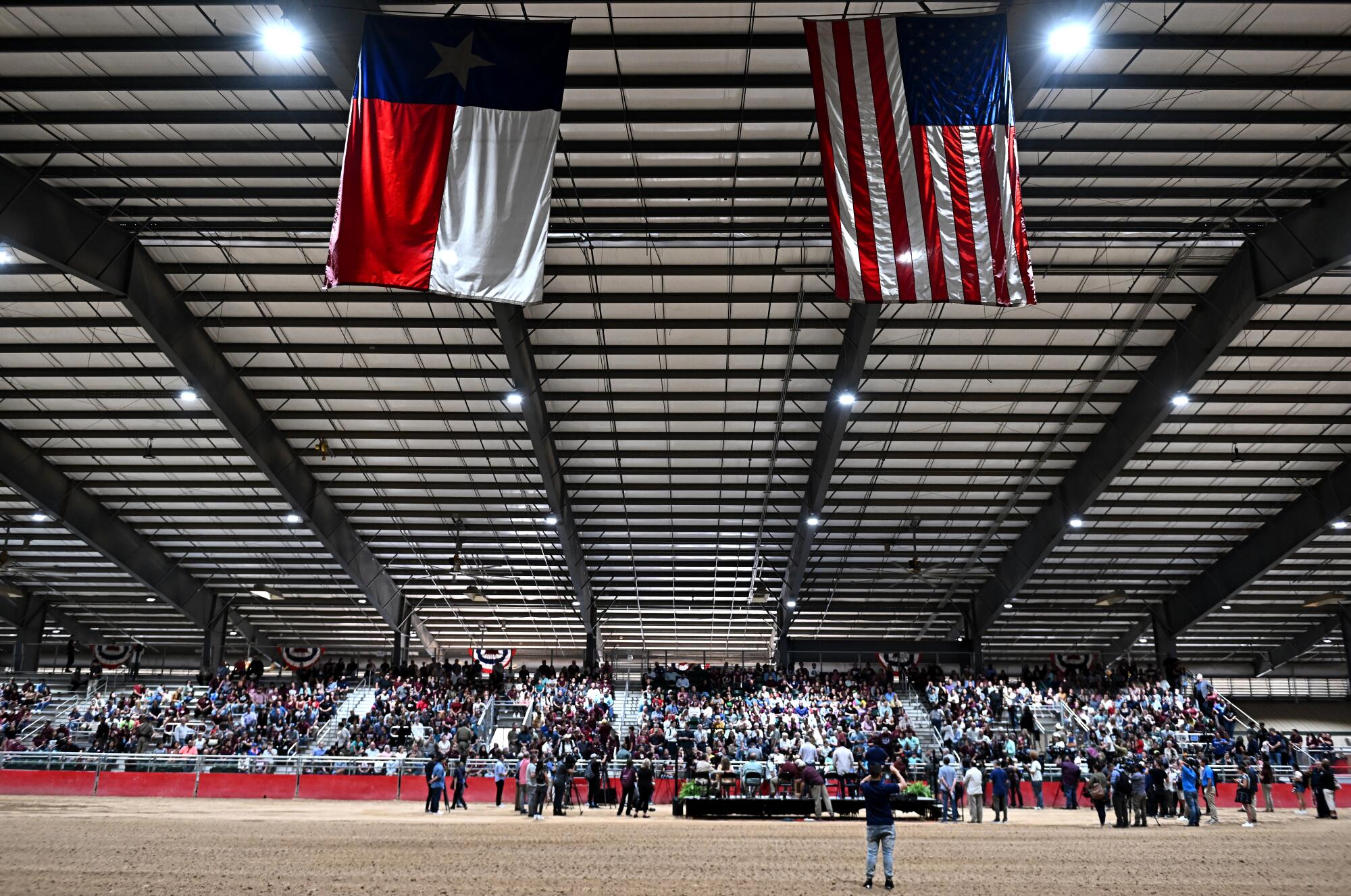 Crowds seated indoors in the bleachers under a blue, red and white flag and the U.S. flag 