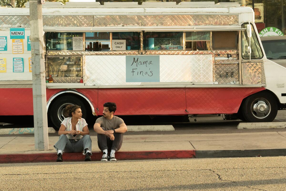 A man and a woman sit on the curb in front of a taco truck labeled "Mama Fina's."