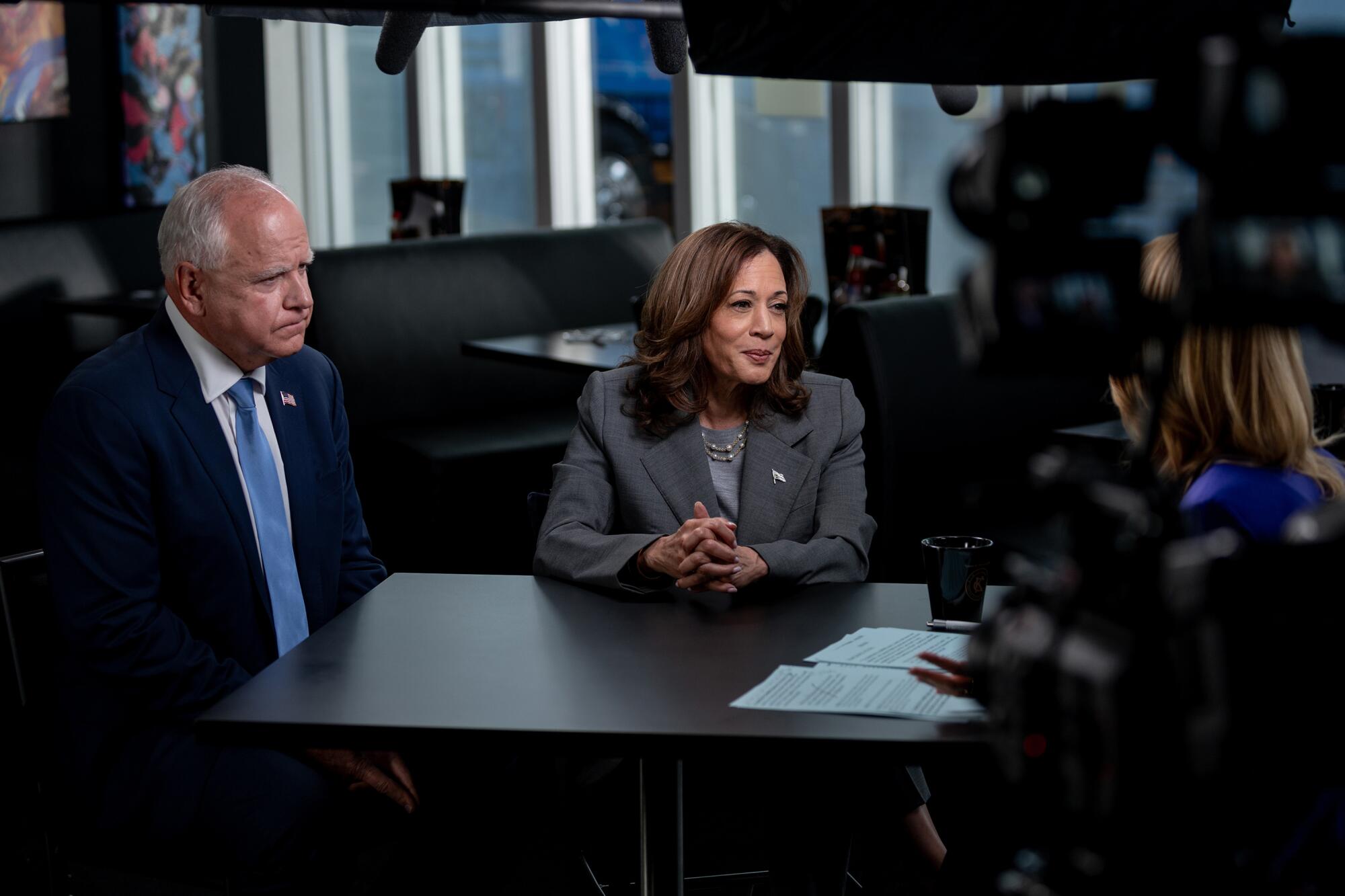 A man in dark suit and blue tie, left, and a woman in a blue suit sit facing another woman 