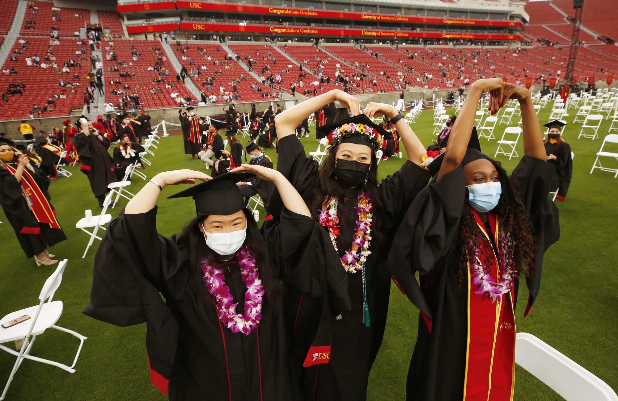 Three students in black graduation gowns arch their arms above their heads to form heart shapes
