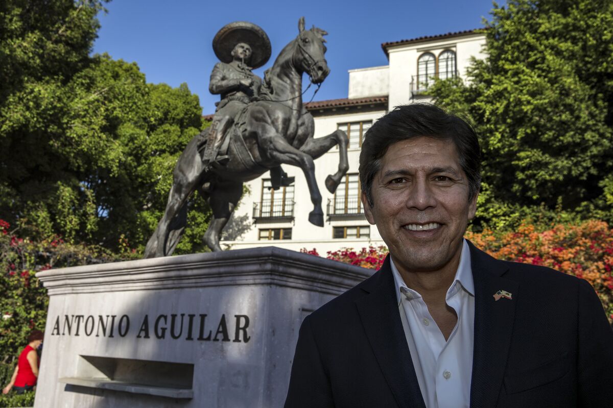 A man in a suit jacket and open collar shirt stands outdoors in front of a statue of a man on a horse.
