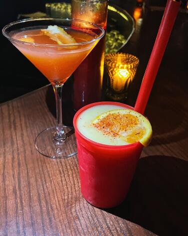 A frozen Orange Julius cocktail with red straw in a red plastic cup next to a pink cocktail in martini glass atop the bar