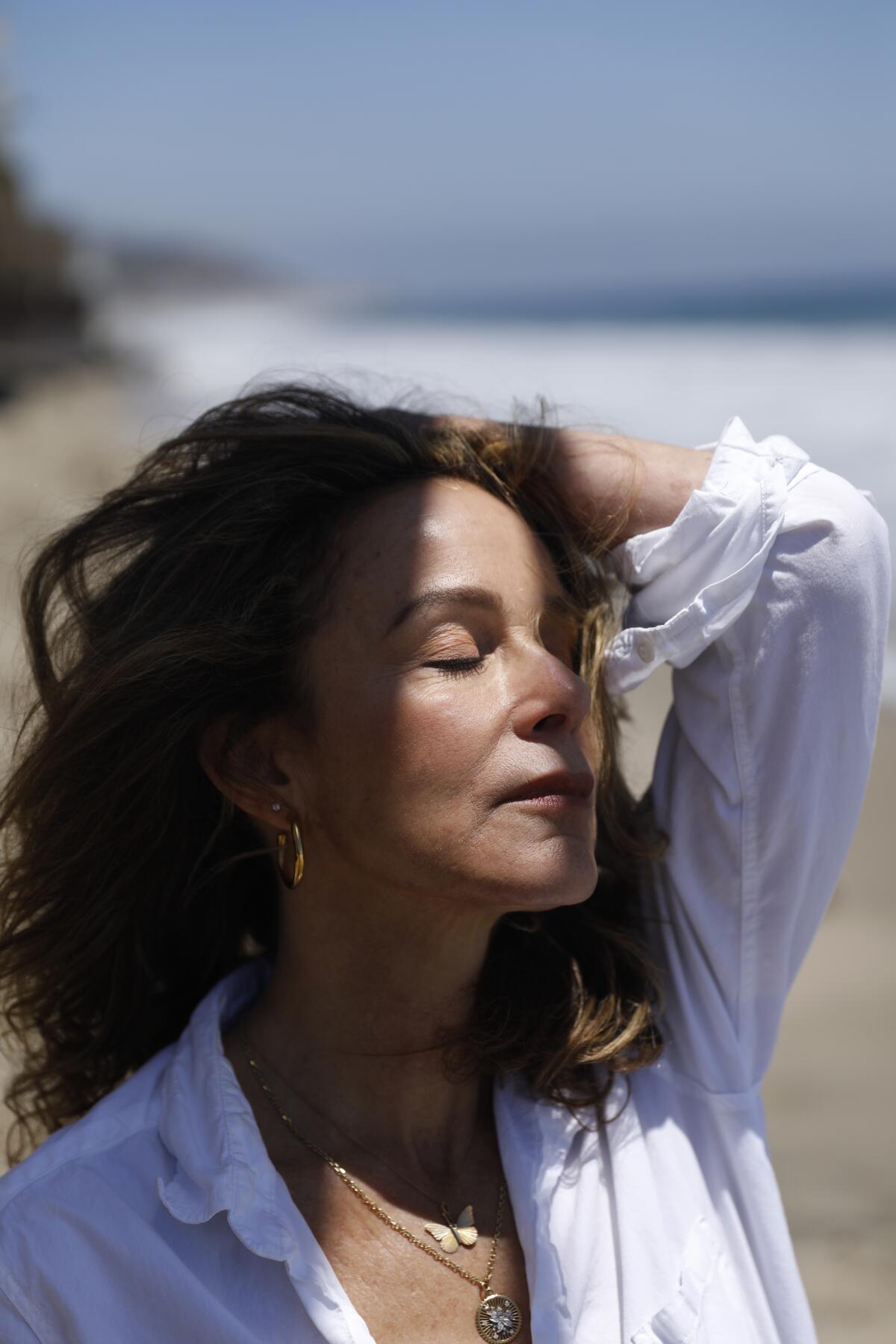 A woman in a white shirt closes her eyes and runs her hand through her hair as sun hits her face