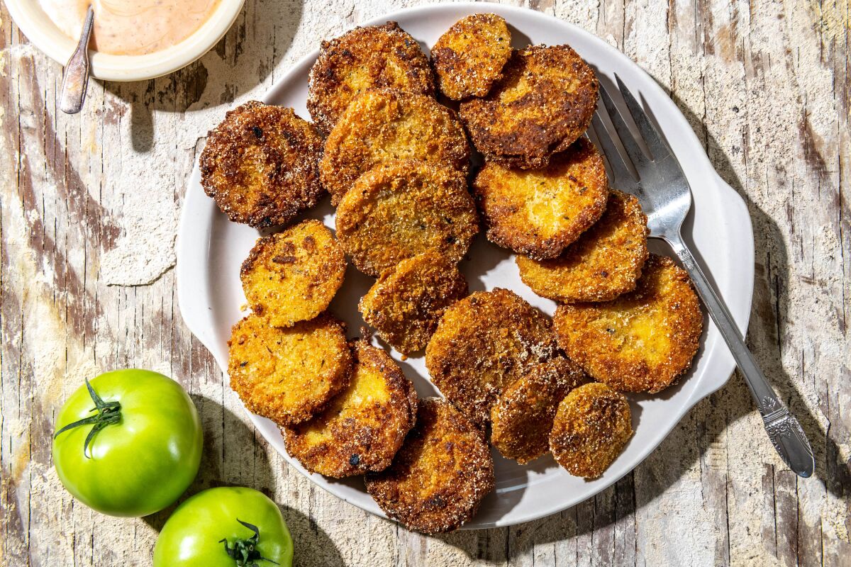 Breaded and fried green tomatoes are seen on a plate, ready to serve, with a few whole green tomatoes nearby.