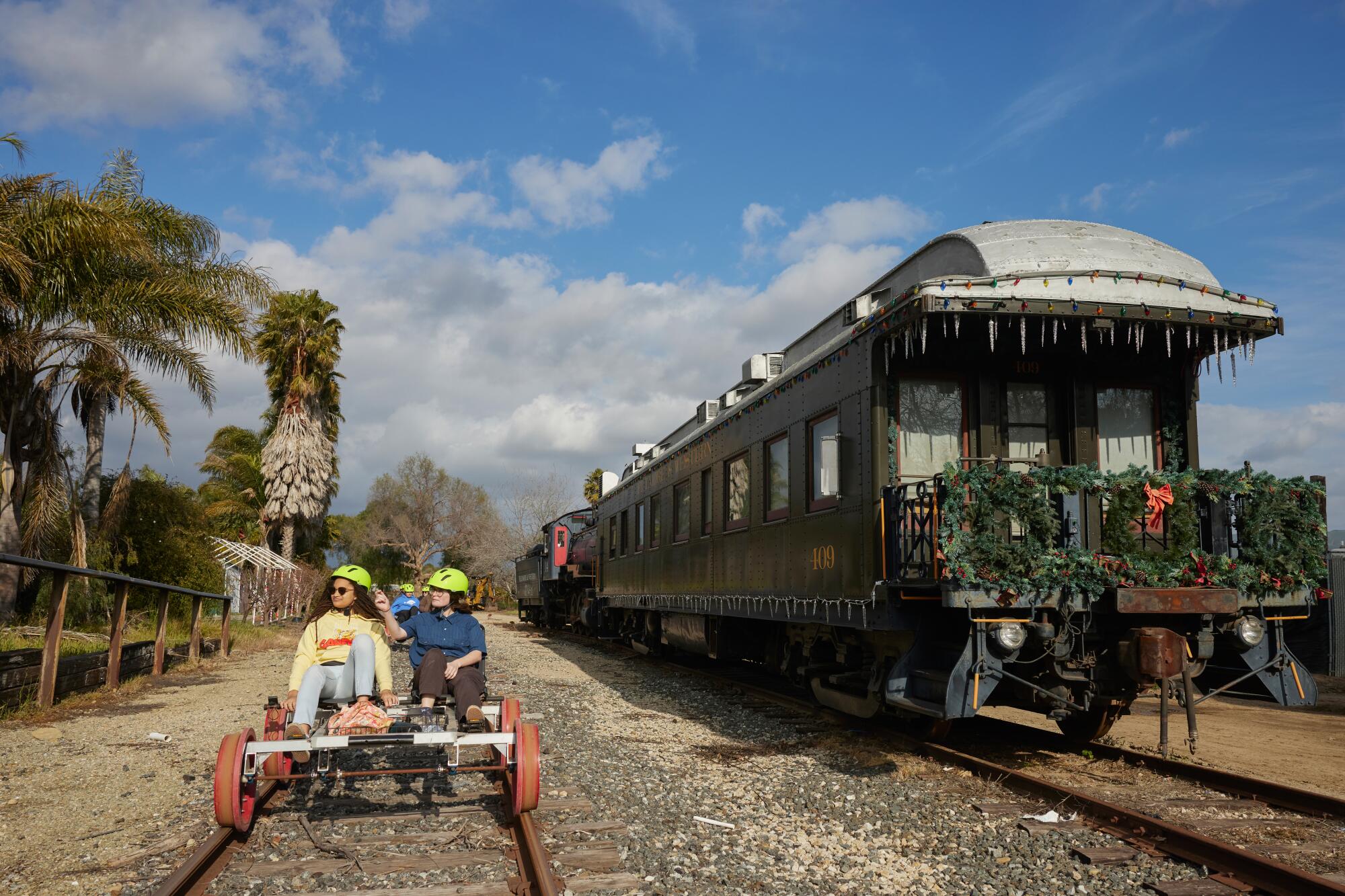 A photo of two people on a railbike, passing by a large train car decorated with lights and wreaths on a sunny day.