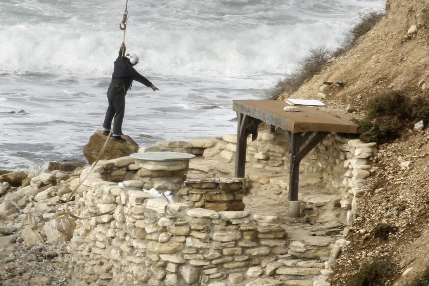 A worker is lowered by a helicopter to start securing heavy equipment to dismantle one of the two surf porches on the shoreline of Lunada Bay.