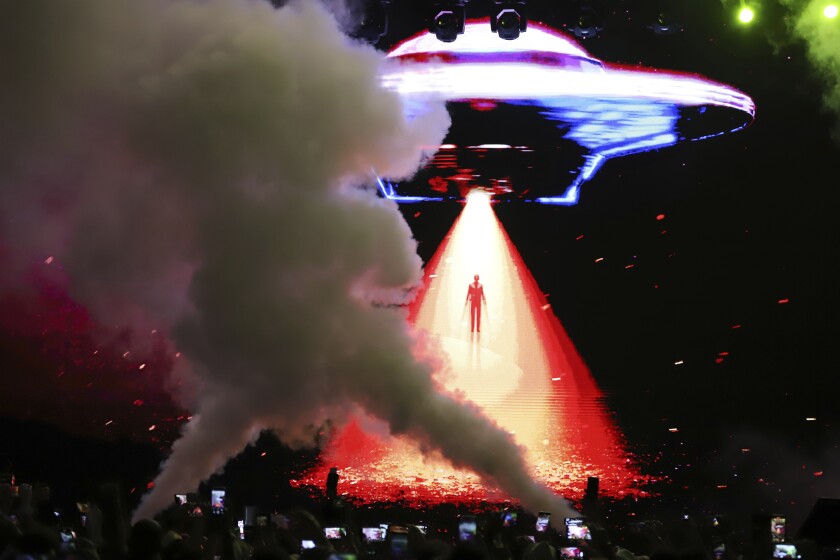 In a dark arena, a crowd of people record on their phones. Above is the outline of a UFO with a red beam of light.