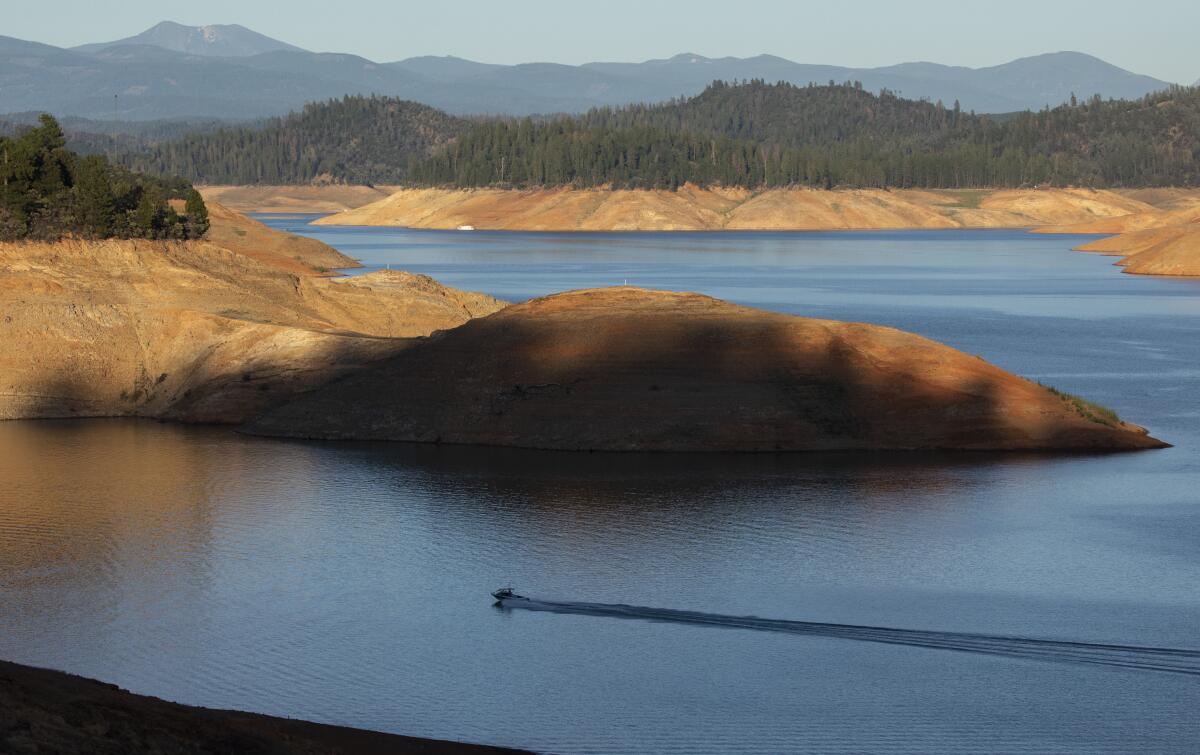 A lake is seen with high, dry banks. In the distance are trees and mountains.