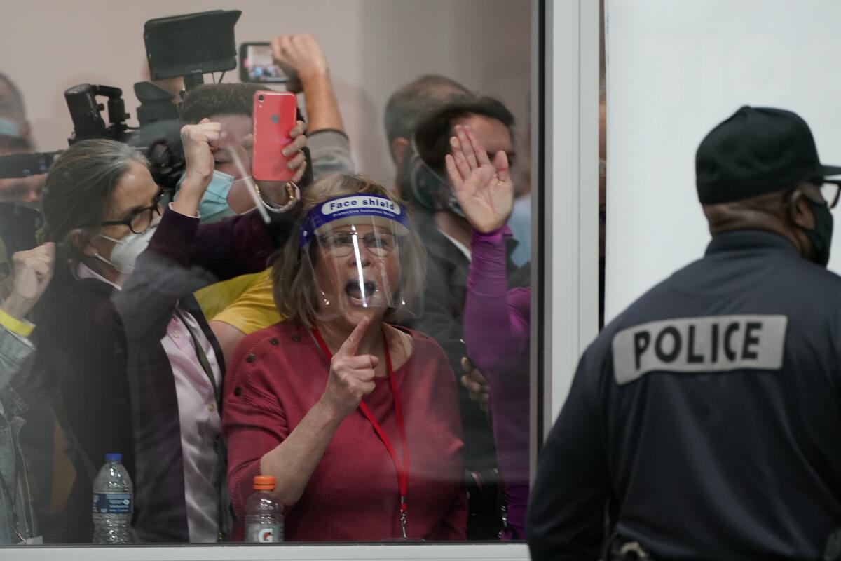 A man in a jacket labeled "Police" stands near a window, behind which people yell and press their hands to the glass.