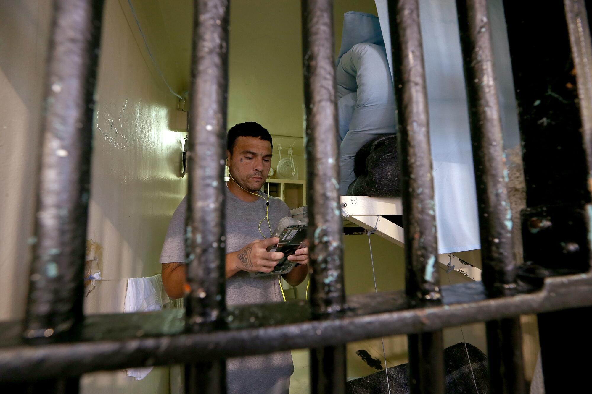 A man standing in a prison cell using a tablet device with earphones, pictured through the metal bars of the cell