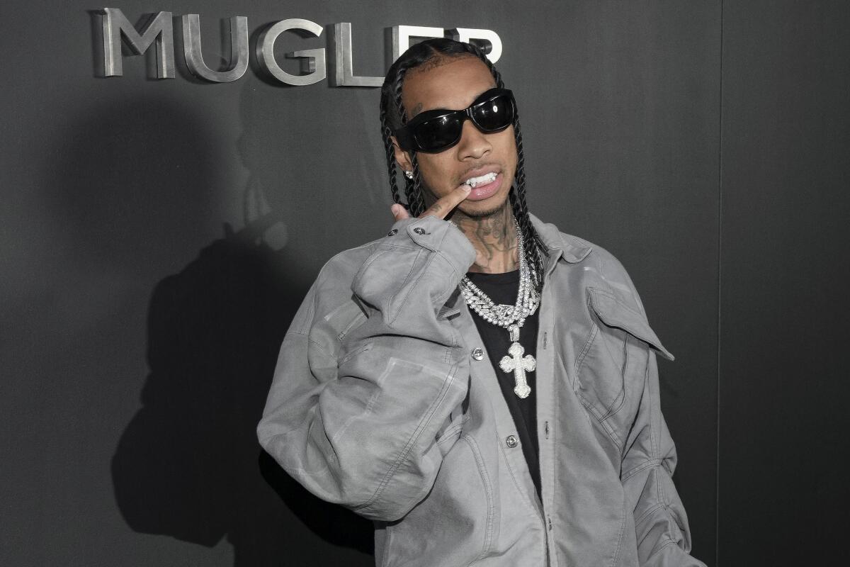 Rapper Tyga wars a gray jacket and black sunglasses as he points to his teeth while posing on a red carpet