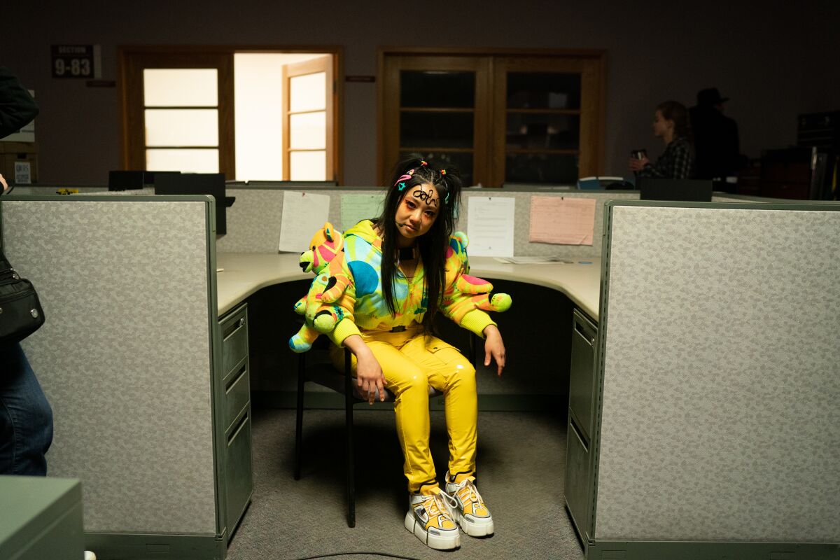 A woman wears bright yellow pants and a multicolored top with stuffed animals attached in a scene from the film.