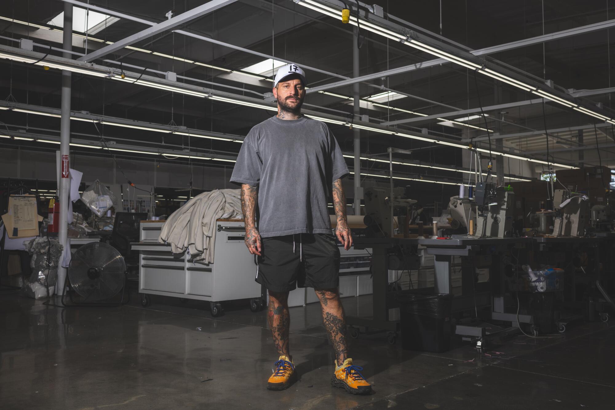 A man stands inside a factory wearing a baseball cap, T-shirt, shorts and sneakers. 