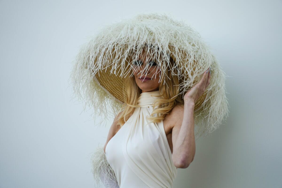 A woman in a clingy white halter dress peers out from under a massive hat covered with white fringe. 
