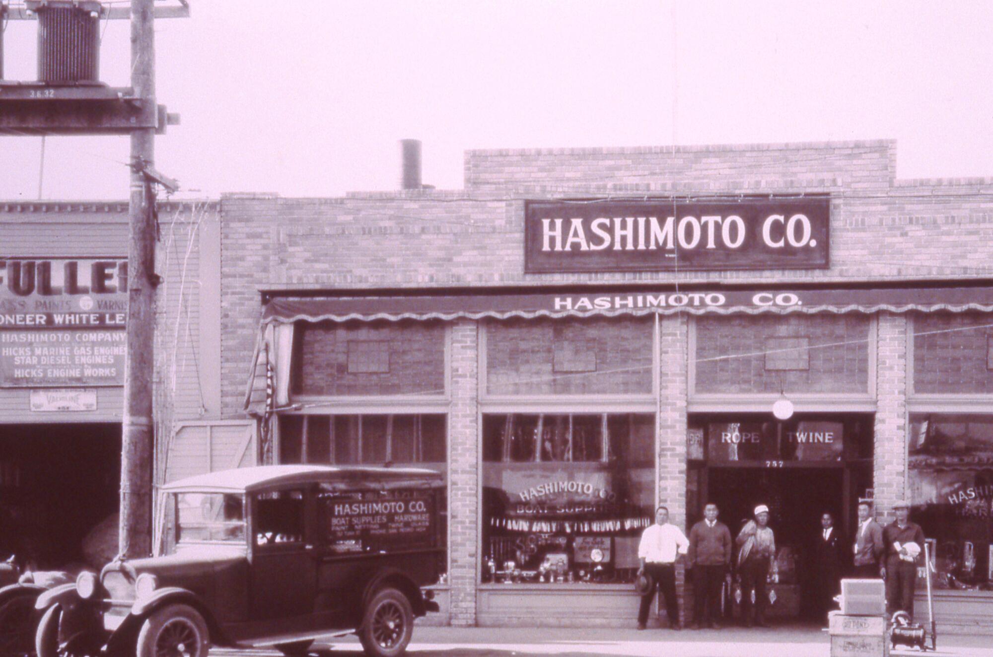 A vintage photo shows a small group of people standing outside a two-story-tall building with a sign that says Hashimoto Co.