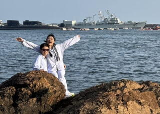 Two women wearing white, one with arms outstretched, are shown near rocks, with the sea and a ship in the background