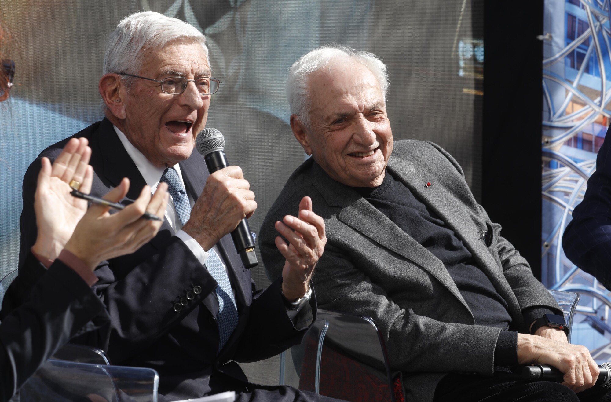 Sitting on a stage with Frank Gehry, Eli Broad speaks into a microphone.