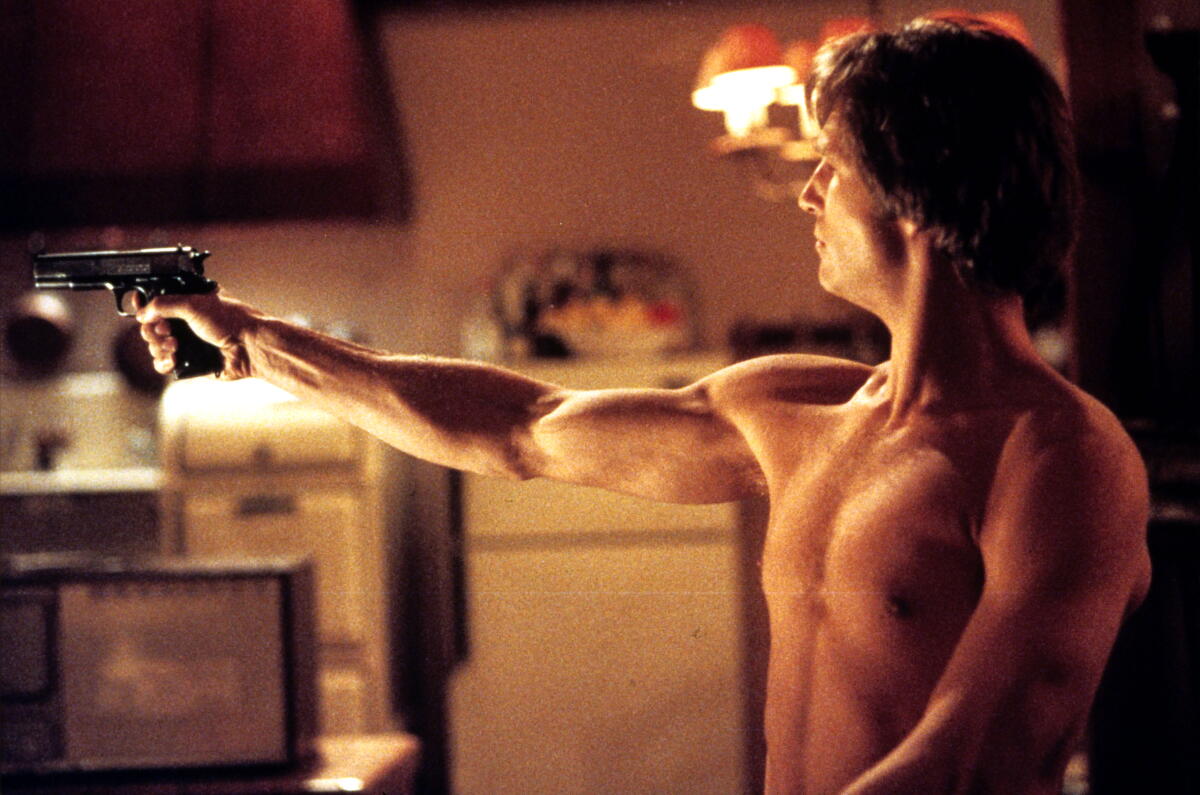 A shirtless man holding up a gun in a kitchen.