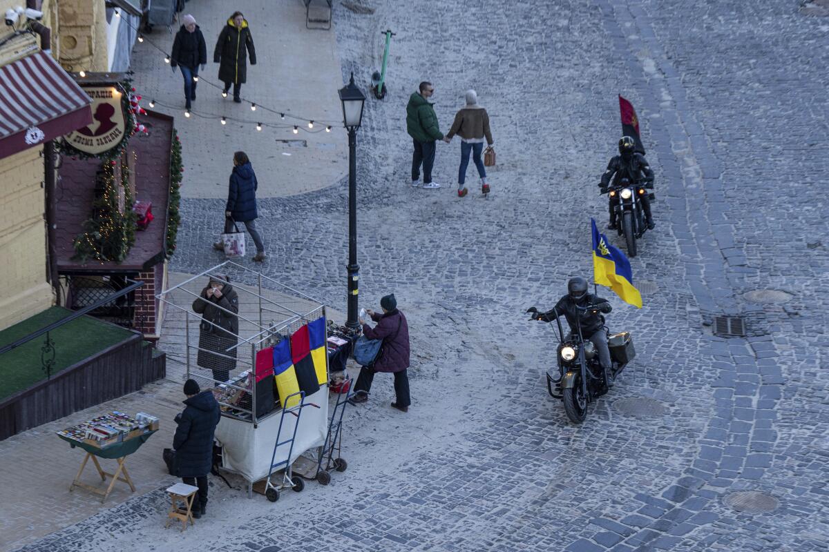 Motorcyclists with blue-and-yellow and red-and-blue flags ride on a snow-covered street near pedestrians 