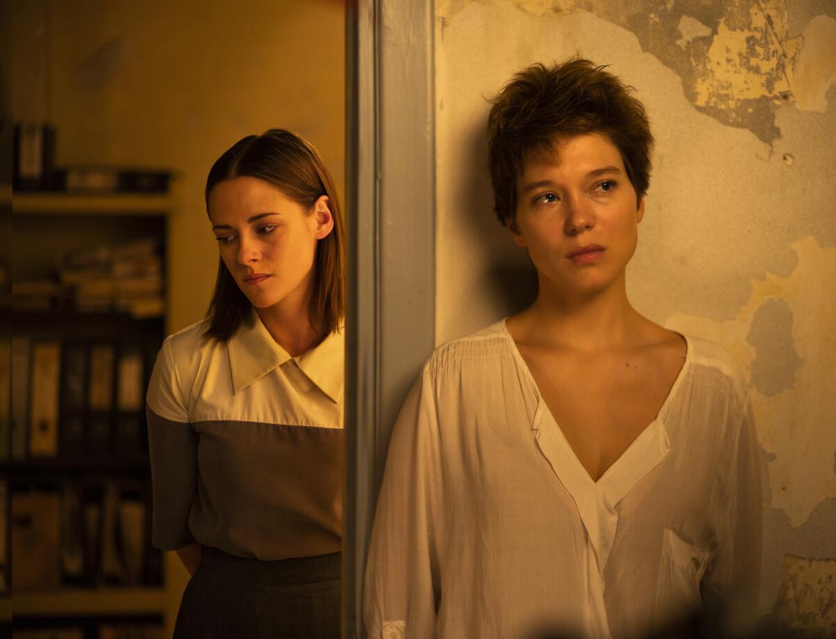 Two young women stand on either side of a door frame, pensively not looking at each other.