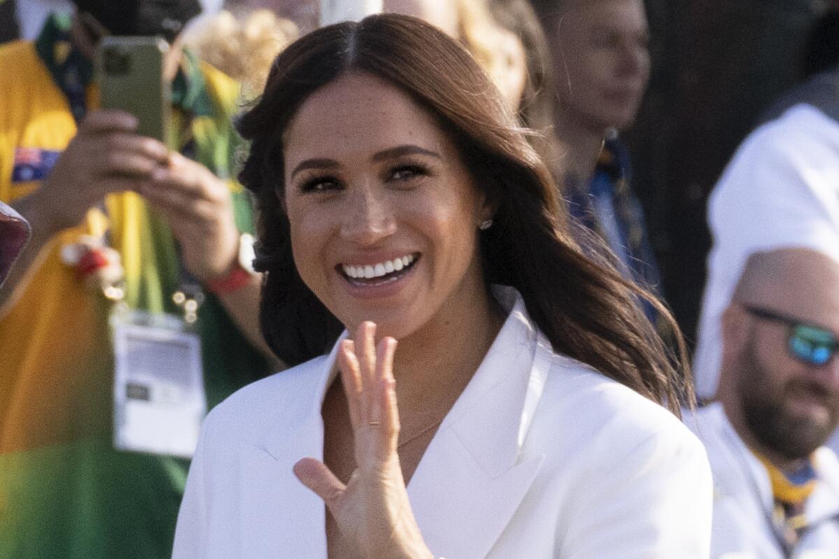 Meghan Markle, in a white collared shirt, smiles and waves her left hand with a crowd behind her.