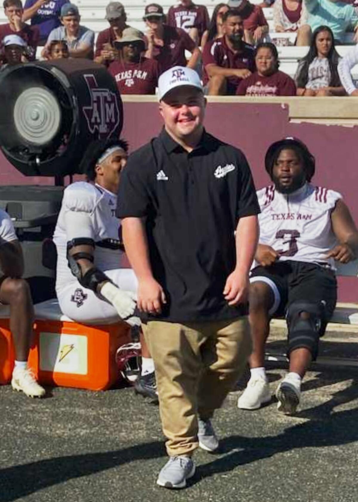 In a vertical frame, Caden Cox poses for a photo standing on a football field with players and fans in stands behind him.
