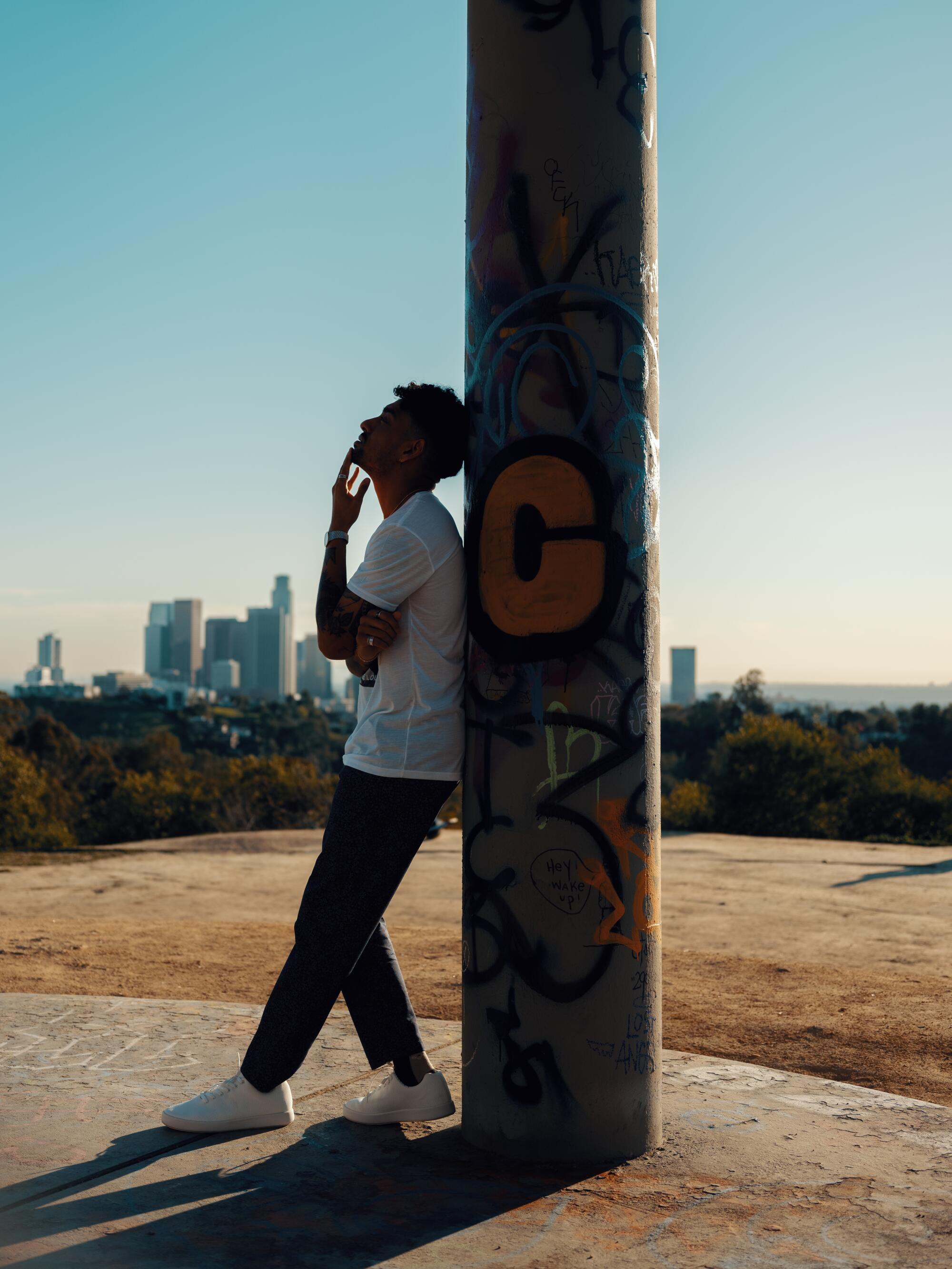 A man leans against a pole covered in graffiti against a view of downtown Los Angeles.