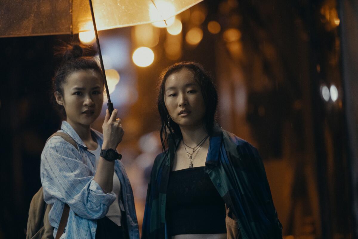 Two women, one holding an umbrella, walk outside on a rainy night in "Expats."