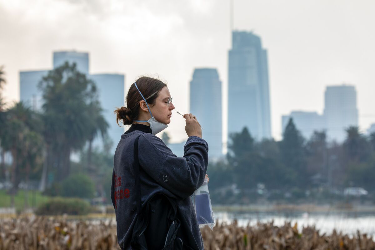 A woman administers a COVID-19 self test with the downtown L.A. skyline behind her.