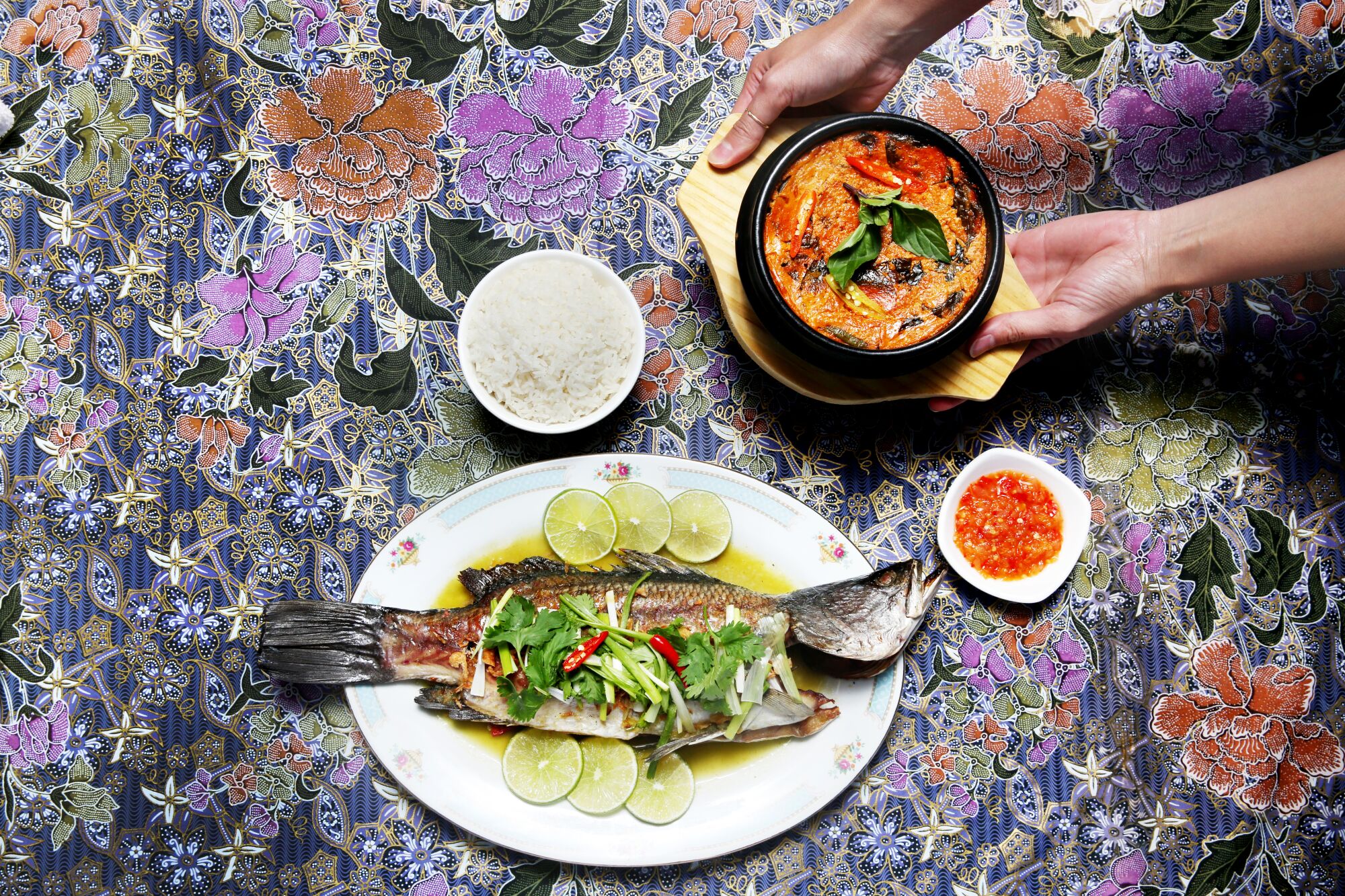 An overhead photo of whole dry-aged fish on a plate, with a set of hands holding a dish of fish curry "custard."