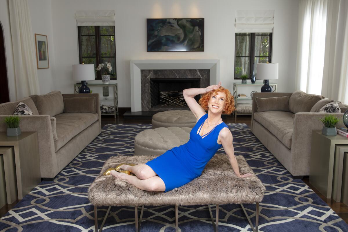 A woman with red hair wears a blue dress and perches on a sofa