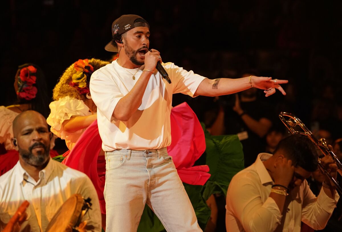 A man in a white T-shirt, jeans and a backward hat sings into a microphone amid a crowd of people with instruments.