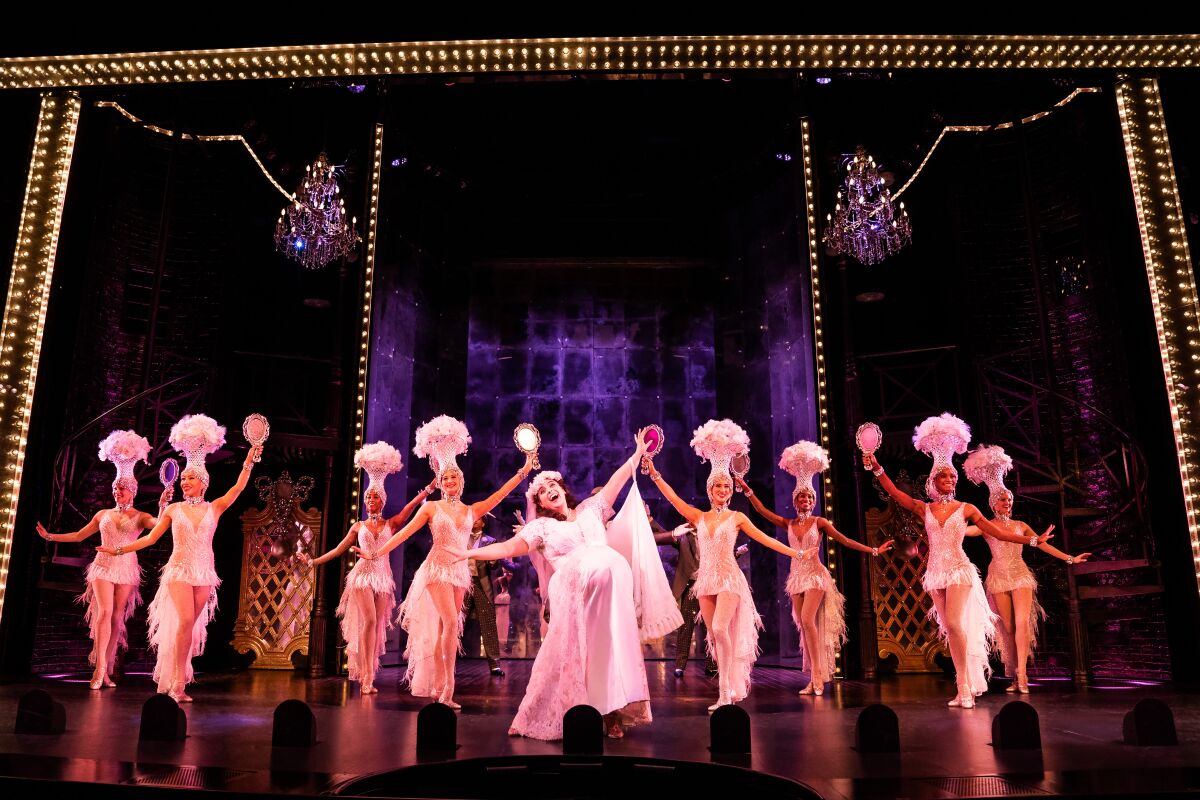 A woman dressed as a pregnant bride in front of a chorus line of dancers in white costumes onstage.